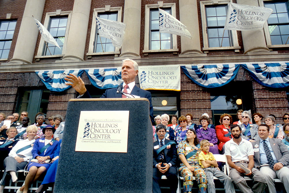 Senator Fritz Hollings speaks at the dedication ceremony for the Hollings Oncology Center in October 1992 with a large crowd seated behind him and flags and bunting on the building