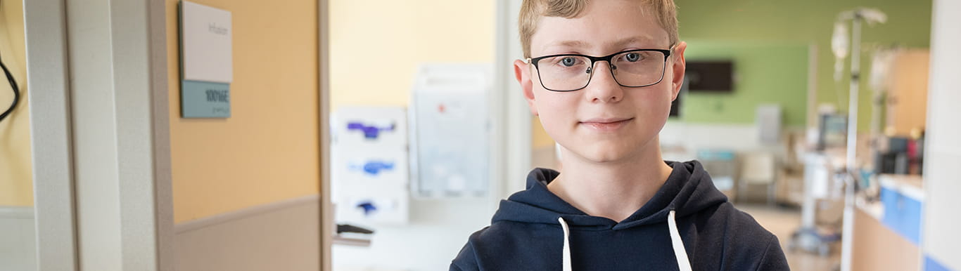 a teenage boy wearing glasses and a navy blue hoodie stands in a hospital corridor