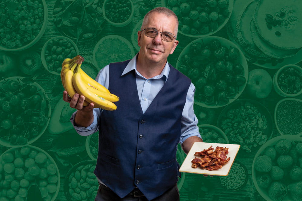 Dr. David Turner holds a bunch of bananas in one hand and a plate of bacon in another with a background of fruits and vegetables