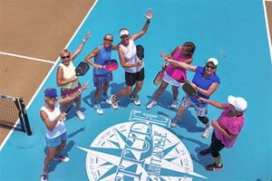 overhead shot of a group of women standing on a pickleball court all pointing to one woman in the middle