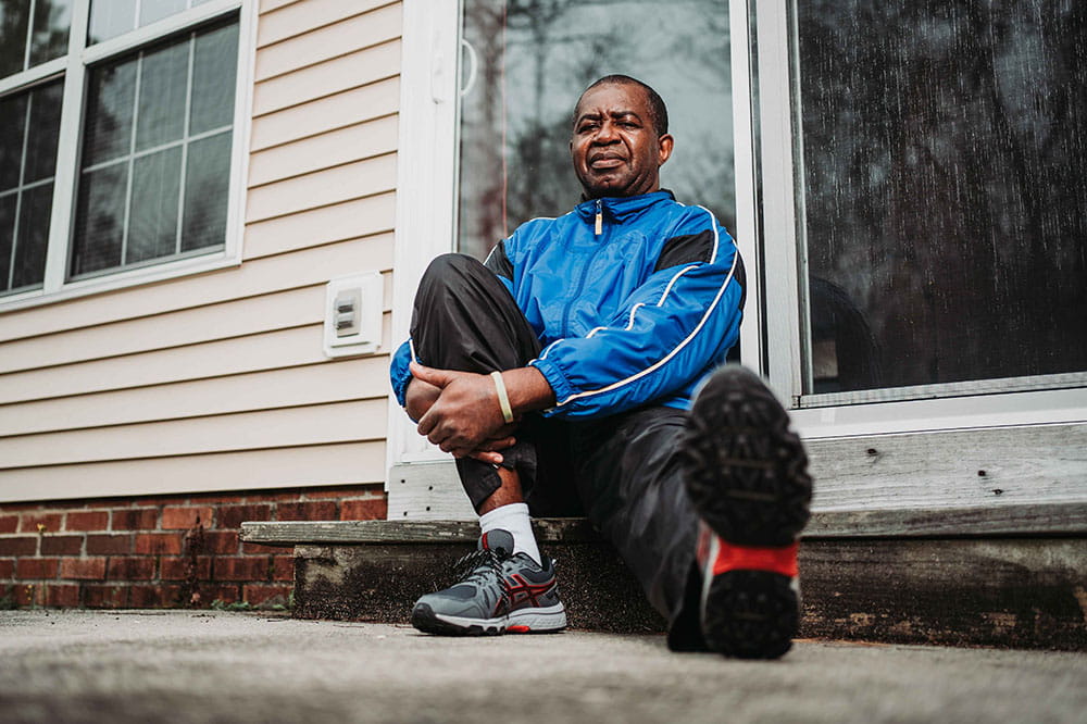 Lee Moultrie sits in front of his house with his arms wrapped around one knee and the other leg extended