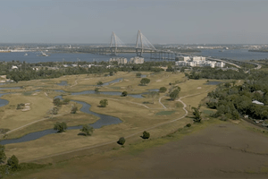 screenshot of an aerial view of the ravenel bridge over the cooper river in the distance with a golf course in the foreground
