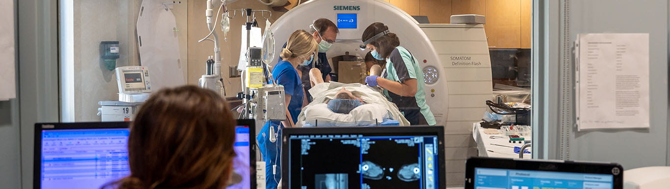 three providers prepare a patient for radiation therapy in treatment room while another provider looks at computer screens