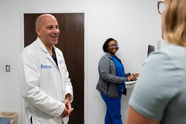 a doctor in a white coat in an exam room chats with a patient while a nurse working at a computer station looks over and smiles