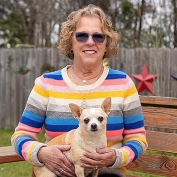 a woman wearing sunglasses sits outside on a bench with a small dog in her lap