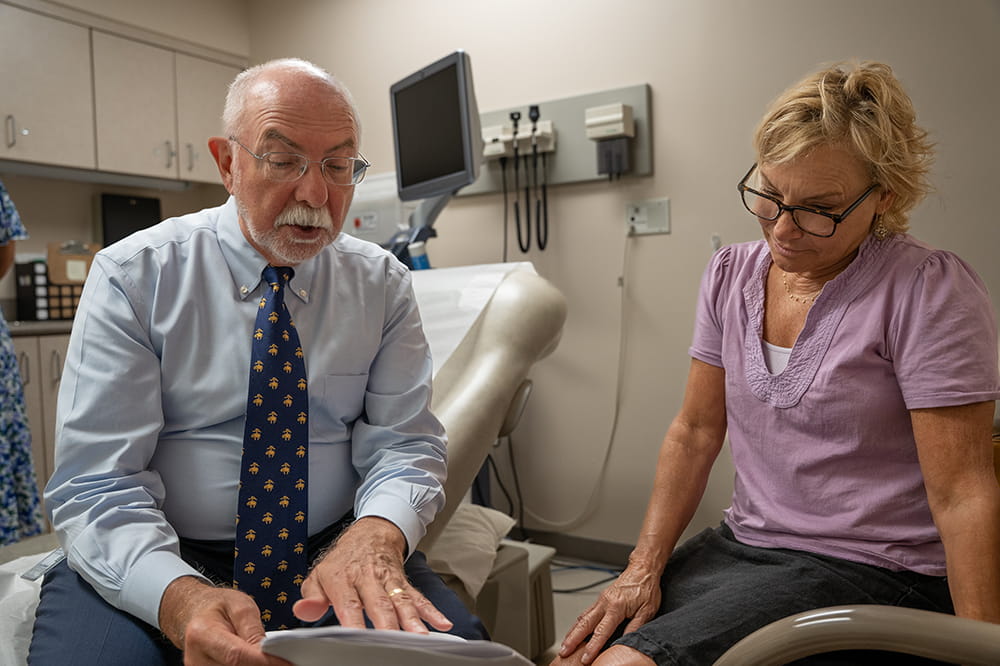 a male doctor in shirt and tie sits and gestures at a paper while talking to a female patient who is also looking at the paper