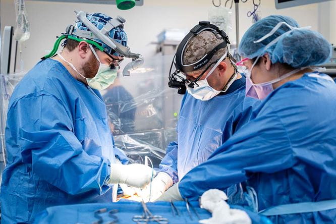 three people in blue surgical gowns and masks lean over someone in the operating room