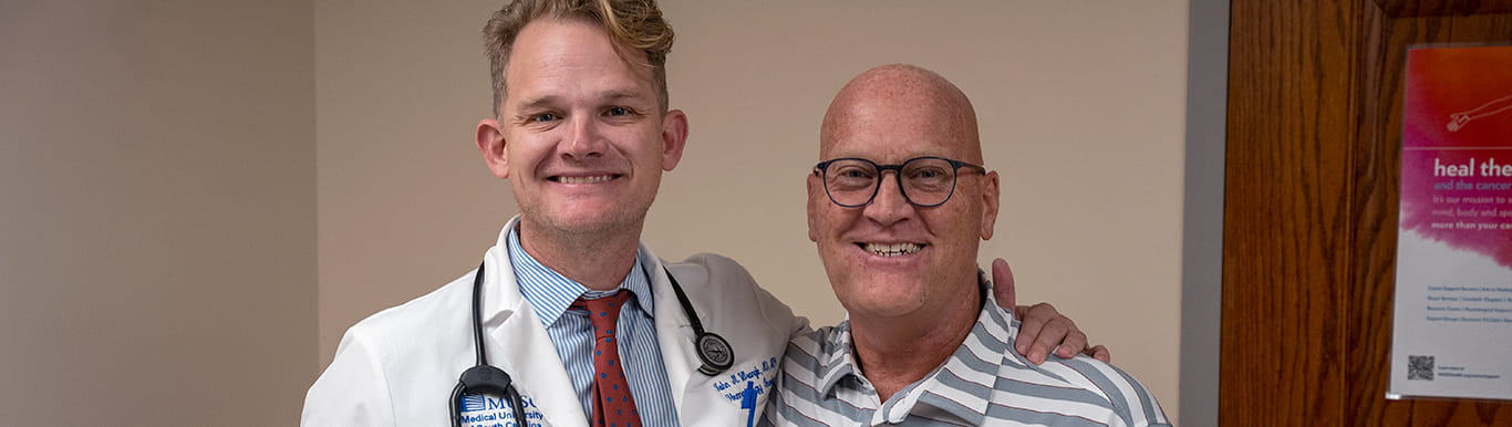 a smiling man wearing a white doctor's coat stands with his arm around another smiling man in a striped shirt