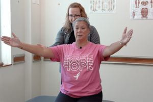 a woman in pink shirt lifts her arms to the side while a physical therapist stands behind her
