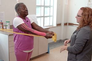 a woman in pink workout clothes uses yellow stretchy bands  for arm strengthening while a physical therapist supervises