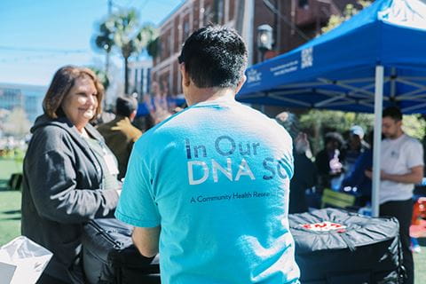 photo focuses on the back of a man's T shirt that says In Our DNA SC as he talks to a woman at an outdoor event