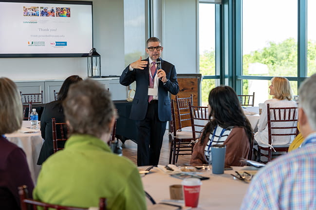 Dr. Frank Penedo holds a microphone and speaks while standing among tables of people at the Scor Symposium