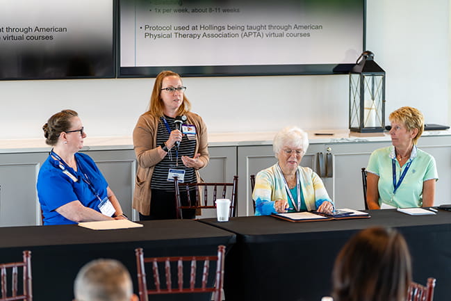 Dr. Katie Schmitt stands and holds a microphone to speak with three other panelists seated next to her at the Scor Symposium