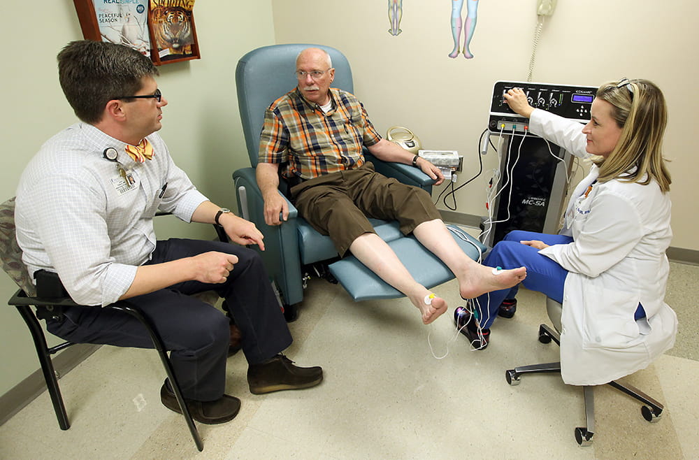 An older man sits in a chair, shoes and socks off and pants rolled up, as a doctor and nurse chat with him