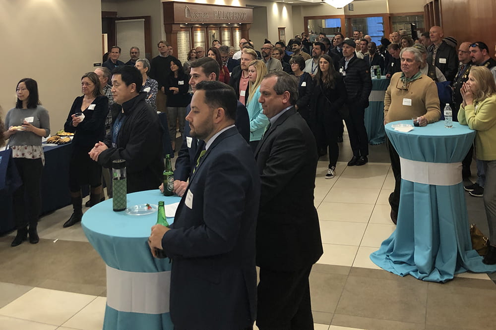 A crowd of people stand and listens to a speaker. 