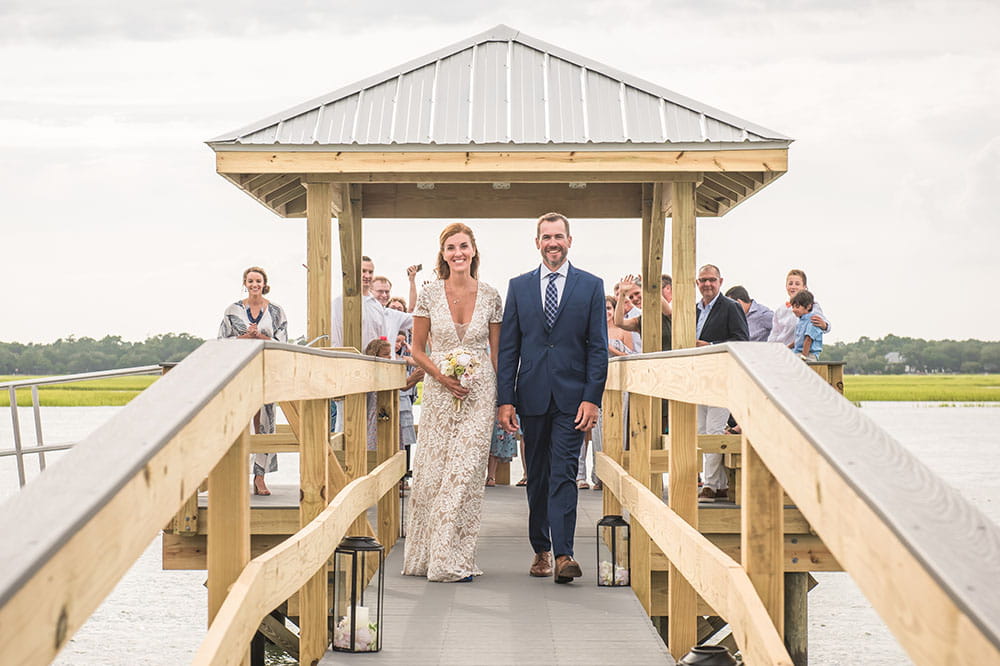 Lara Lambert and Carson Thomas walk down a dock after their wedding ceremony with people looking on behind them