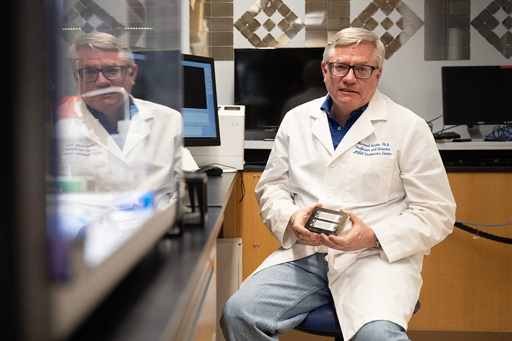 Richard Drake sits in his lab and holds a component of the GlycoTyper technology