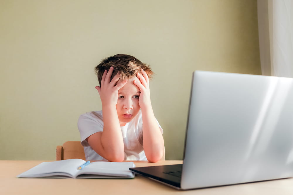 A boy with his hands on his face as he stares in frustration at a computer screen.