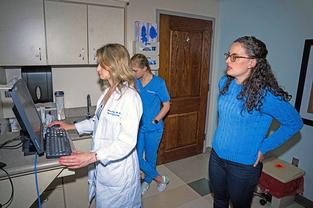 in an exam room, a doctor stands and looks at a computer screen while a patient stands slightly behind her also looking at the screen 