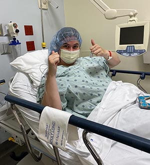 a woman in a hospital bed with a mask and hair net gives a double thumbs up