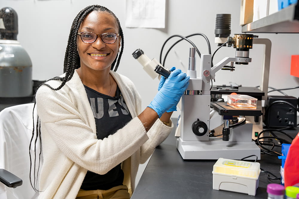 a young woman sitting in front of a microscope looks at the camera and smiles