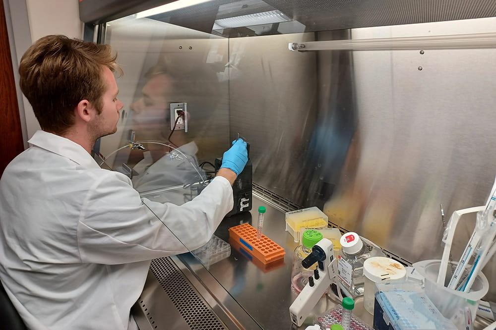 a man in white coat and blue gloves reaches under a glass hood to carefully test the hydropore device