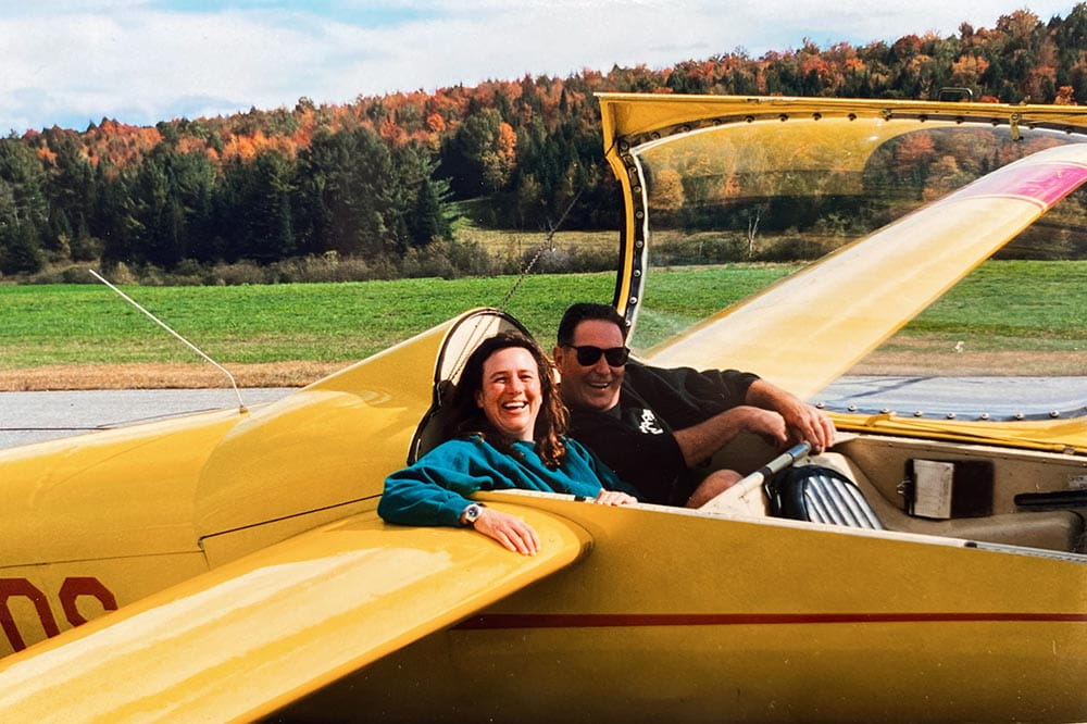In a photo of indeterminate vintage, a man and a woman sit in a yellow biplane on a runway with a woods of fall colors behind them