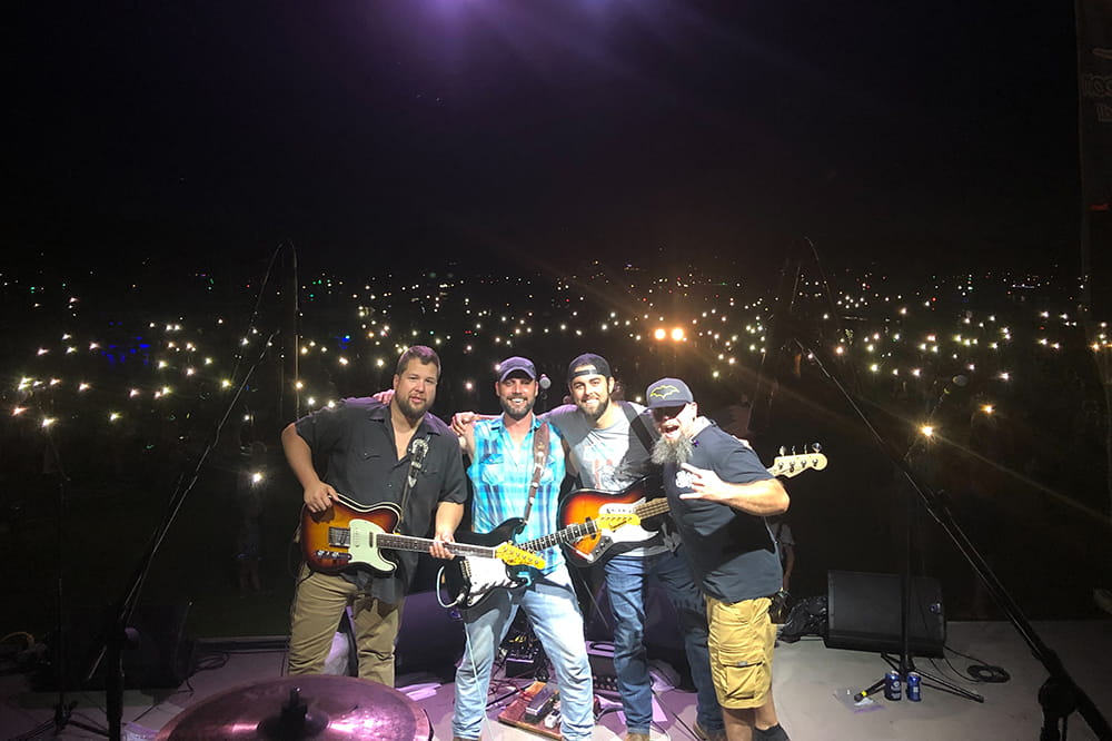 four band members stand on an outdoor stage at night posing for camera with the lights of the city behind them