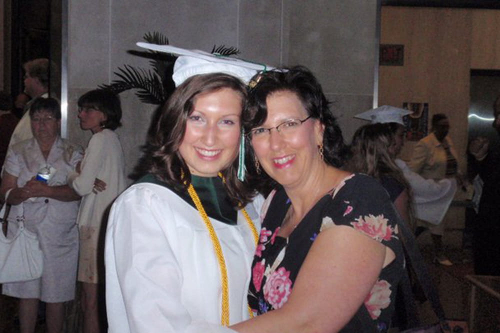 a woman in a white graduation gown and cap poses, hugging her mother