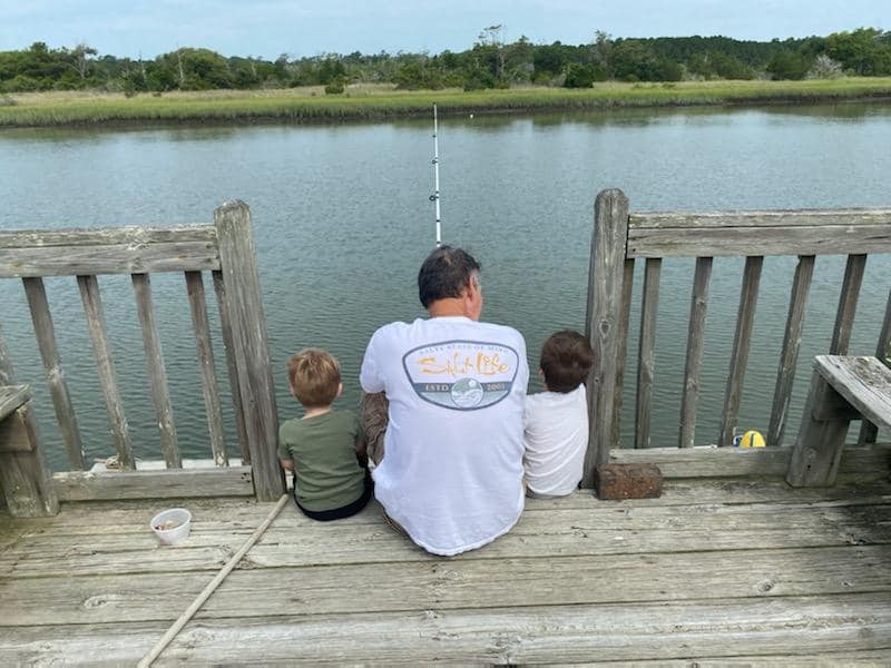 seen from behind, a man sits at a dock fishing with a small boy on either side of him