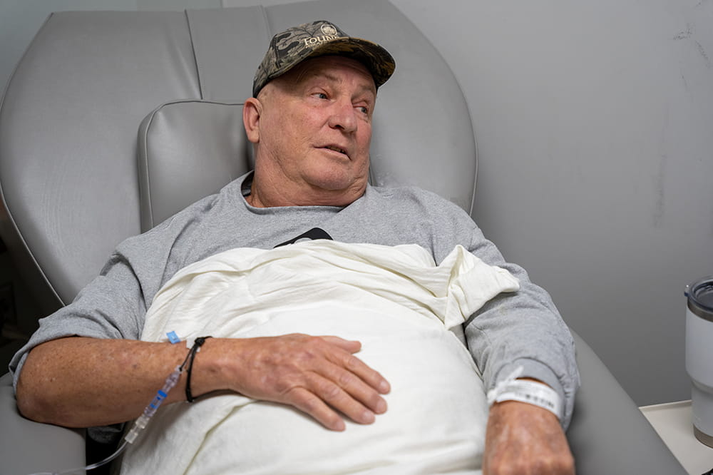 a man in gray sweatshirt with sleeves pushed up and a camo baseball cap sits in a chair with a white blanket draped over him and hospital bracelets on