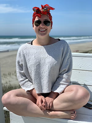 a smiling young woman sits cross legged on a bench at the beach with a bright red scarf on her head