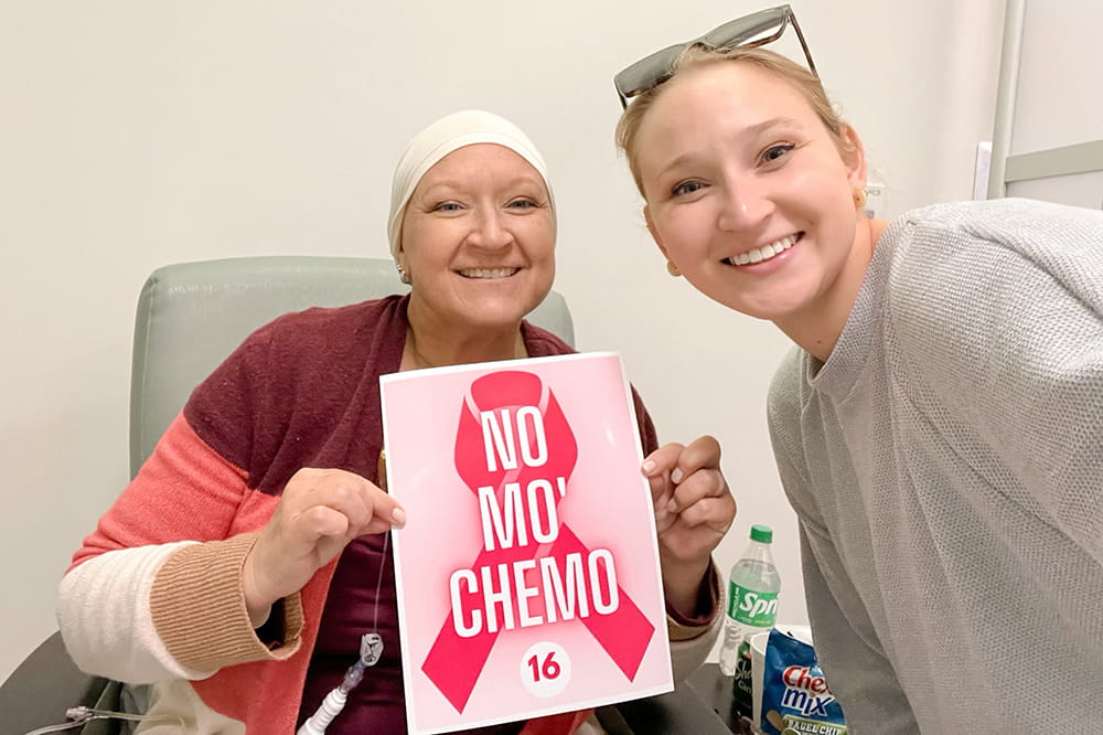 a woman in hospital chair wearing head scarf holds a sign that says No Mo Chemo while a younger woman smiles with her