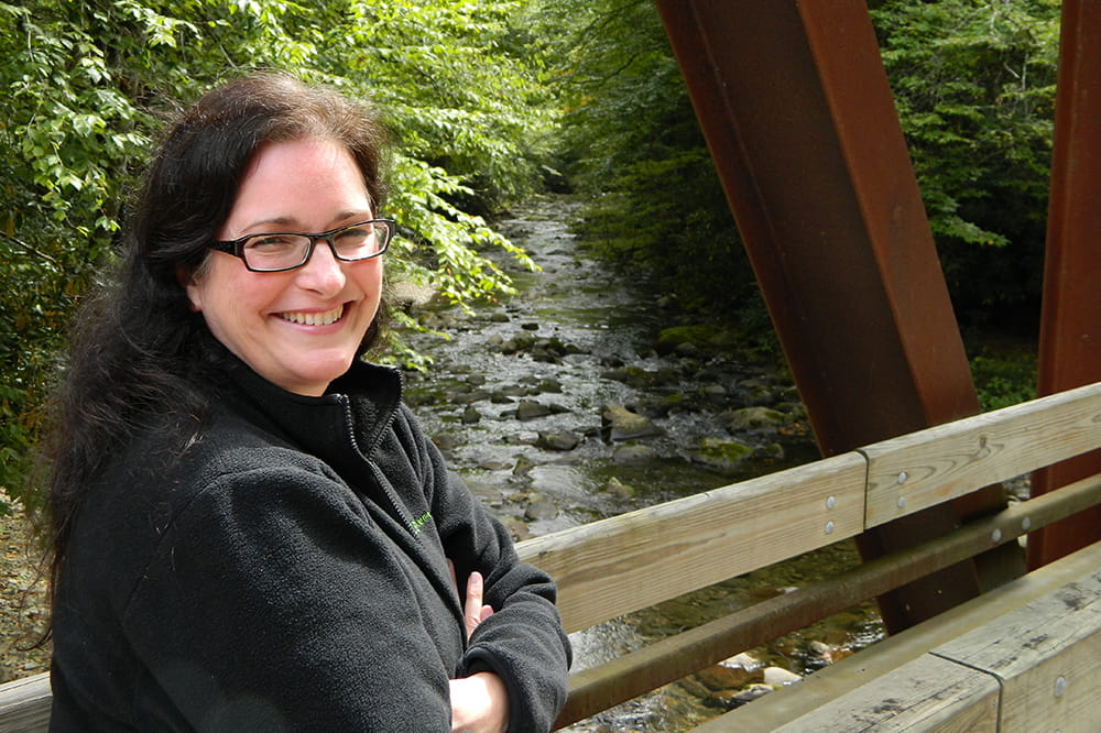  a smiling woman standing on a wooden bridge over a creek looks over her shoulder at the camera