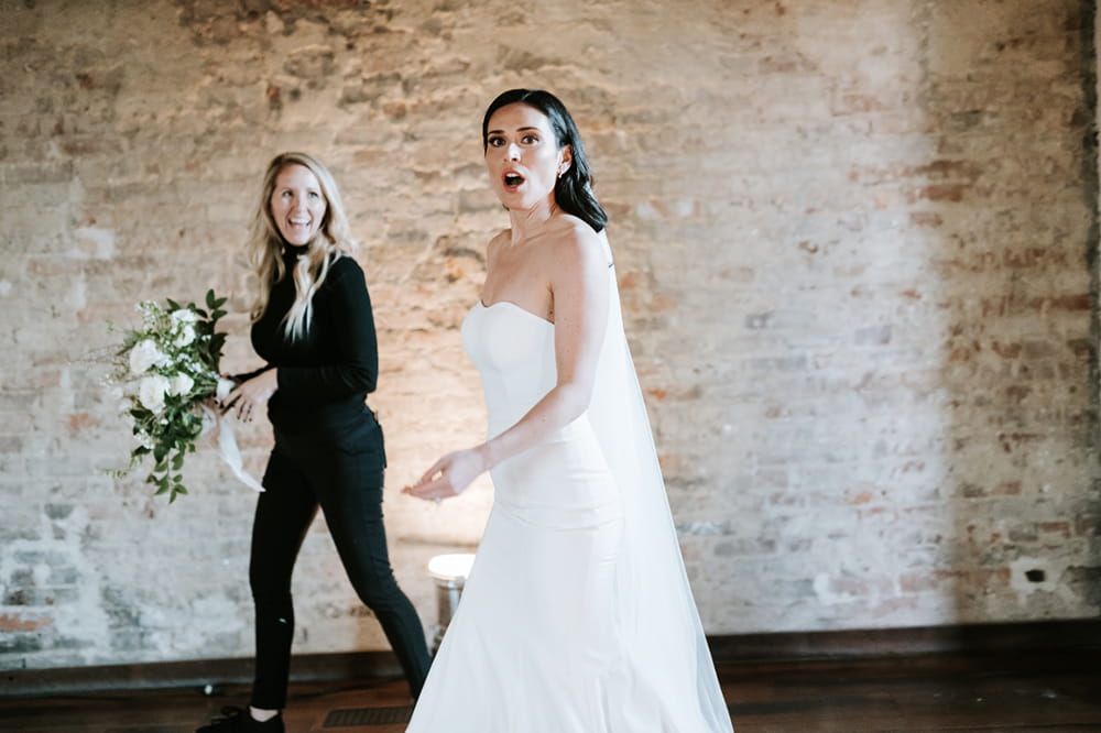a bride looks startled at the camera while a woman all in black in the background smiles affectionately