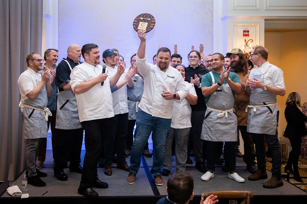 a group of people in chefs uniforms stand on a stage and clap while one in the middle triumphantly holds aloft a plaque with a gold fork attached