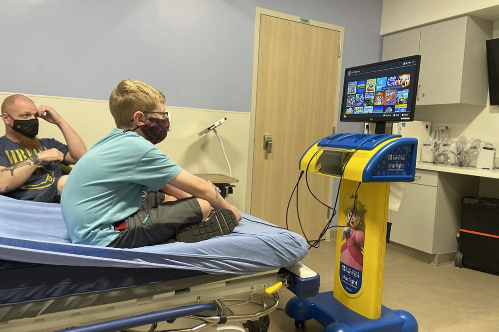 a boy sits cross legged on a hospital bed playing video games on a screen that sits on a tall podium like stand that has been rolled into the room