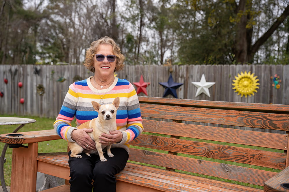 a woman sits on a wooden bench in her backyard with a Chihuahua on her lap