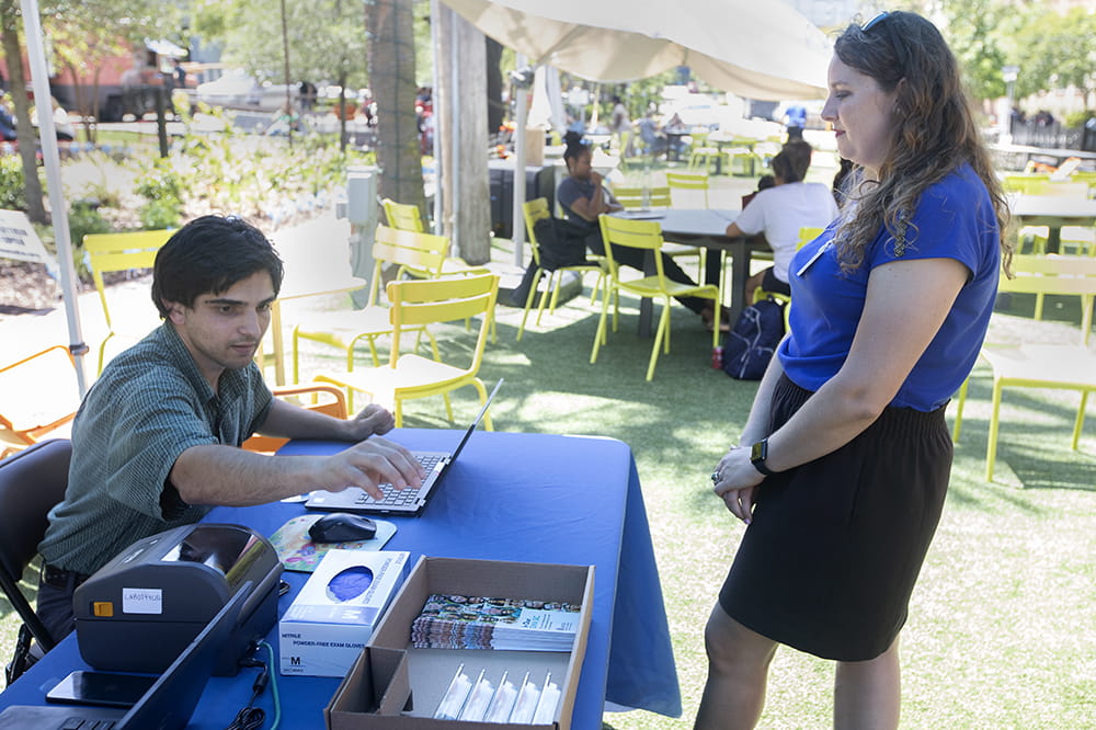 Man seated at a table with a laptop computer looks around while a woman with curly hair stands in front of him.