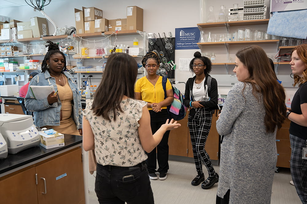 a loose circle of young women stands in a lab listening to one talk