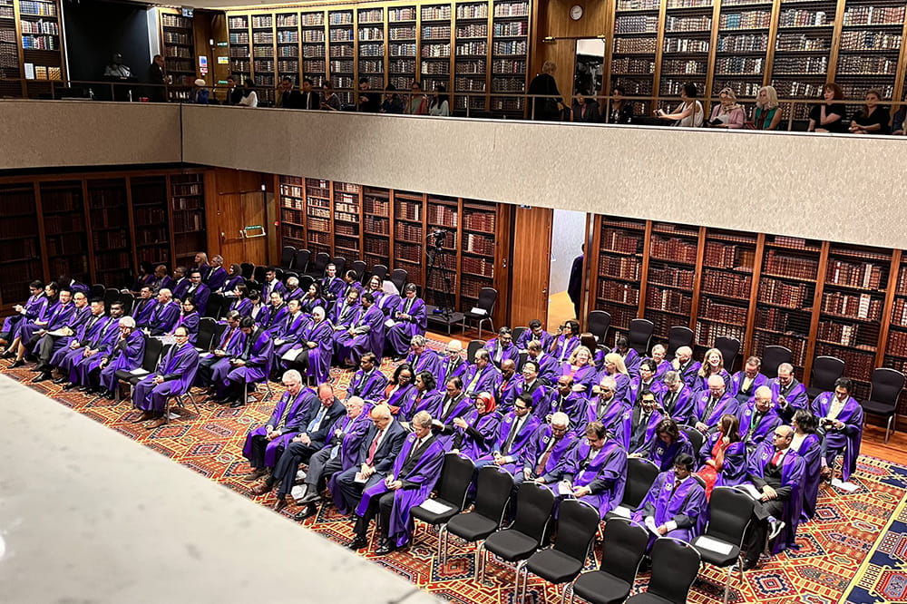 a group of people in purple robes sit in an open area in an elegant old library, while people on balconies circling the open area observe