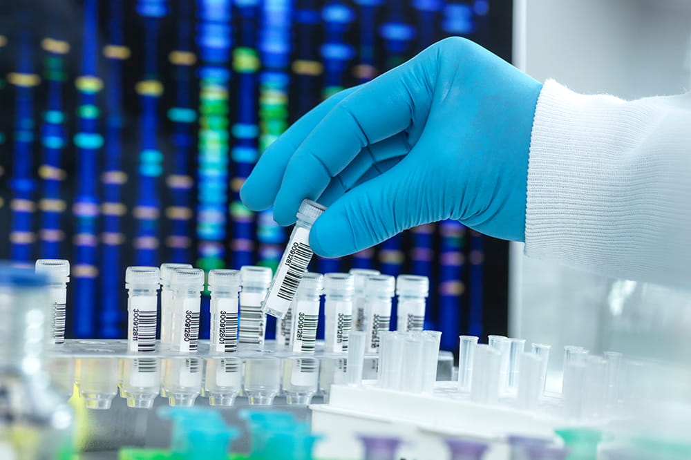 a glove hand picks up a glass vial in front of a computer screen showing rows of DNA data