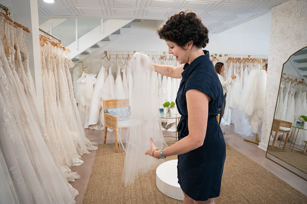 a woman smooths a white veil as she stands in a shop surrounded by wedding dresses