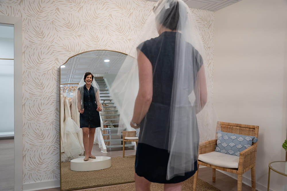 a woman wearing a bridal veil and regular day clothes looks at herself in full length mirror in a bridal shop