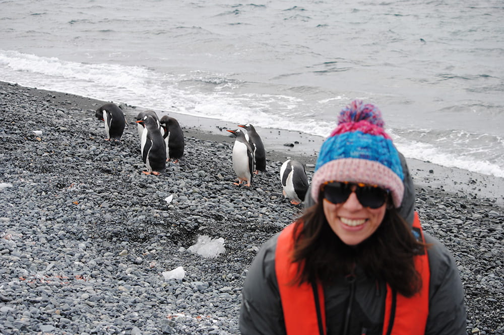 a woman grins at the camera as behind her penguins go about their day