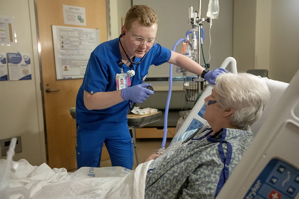 Nurse leans over to talk with a patient who is sitting up in an adjustable bed.