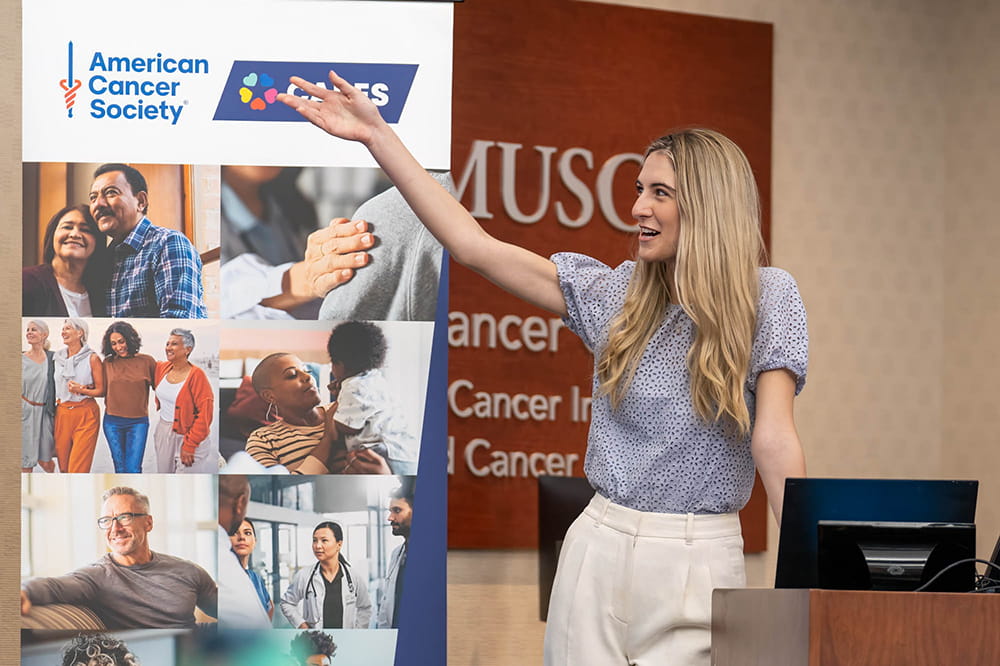 a woman stands at a podium and gestures to an American Cancer Society banner to her side