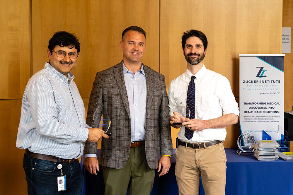 three men in business casual pose with two holding clear  awards