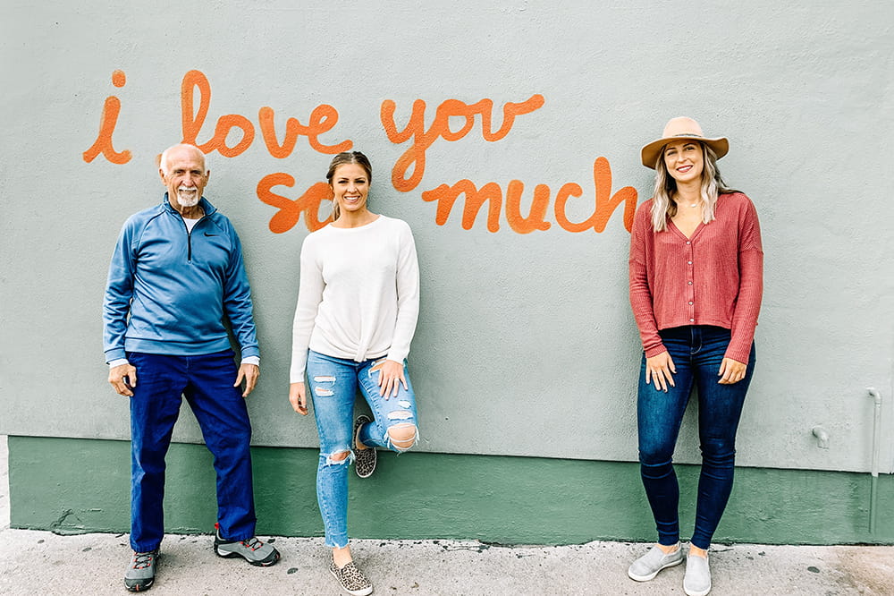 an older man and two young woman pose in front of a wall that says in orange script I love you so much