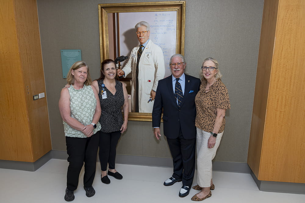 three women in Sunday casual and a man in a suit pose in front of a life-size painting of the man. In the painting he he wearing a white doctor's coat